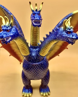 A detailed and vibrant toy dragon with three heads, each featuring sharp teeth and small horns. The body and wings are a striking blue color with glittery scales, paired with golden accents on the horns, wing edges, and necks. The dragon stands with its wings slightly spread on a plain background.