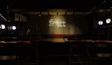 A dimly lit interior of a cafe or restaurant with chairs placed upside down on tables. The setting appears to be closed. A neon sign on the wall reads 'Brewing Stories' and provides a focal point in the dim space, while round lights illuminate some areas.