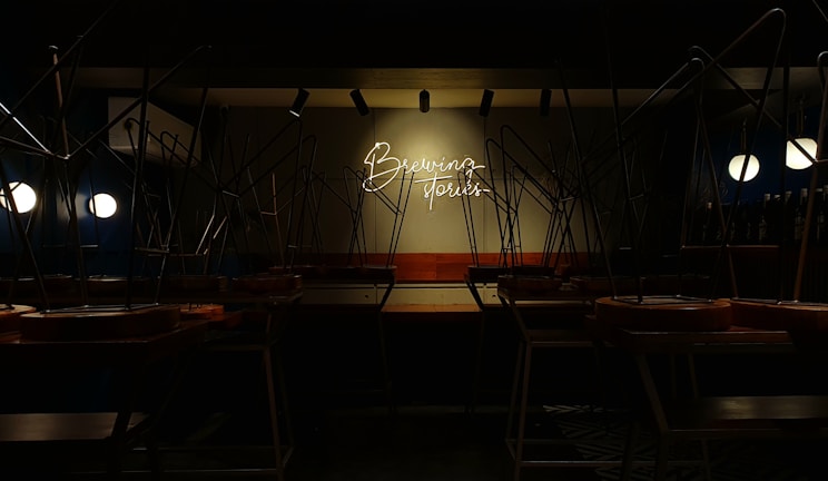 A dimly lit interior of a cafe or restaurant with chairs placed upside down on tables. The setting appears to be closed. A neon sign on the wall reads 'Brewing Stories' and provides a focal point in the dim space, while round lights illuminate some areas.