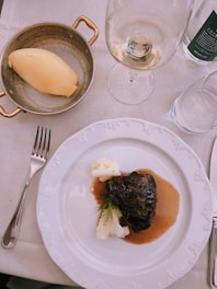 A white plate with a dark piece of braised meat garnished with dill alongside creamy mashed potatoes, all drizzled with a brown sauce. Next to the plate sits a small copper pan containing a scoop of mashed potatoes. A wine glass and a water glass are also visible, all arranged on a white tablecloth.