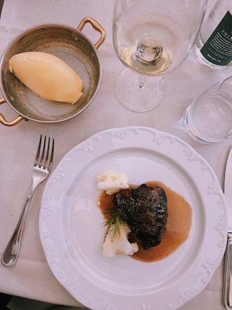 A white plate with a dark piece of braised meat garnished with dill alongside creamy mashed potatoes, all drizzled with a brown sauce. Next to the plate sits a small copper pan containing a scoop of mashed potatoes. A wine glass and a water glass are also visible, all arranged on a white tablecloth.