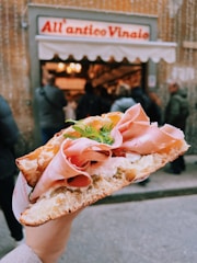 A hand holds a large, freshly made sandwich filled with thinly sliced ham, cheese, and arugula, with a backdrop of a bustling deli entrance and its sign.