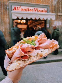 A hand holds a large, freshly made sandwich filled with thinly sliced ham, cheese, and arugula, with a backdrop of a bustling deli entrance and its sign.