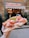 A hand holds a large, freshly made sandwich filled with thinly sliced ham, cheese, and arugula, with a backdrop of a bustling deli entrance and its sign.