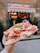 A hand holds a large, freshly made sandwich filled with thinly sliced ham, cheese, and arugula, with a backdrop of a bustling deli entrance and its sign.