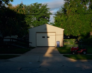 A garage with a closed door is partially bathed in sunlight, creating a stark shadow across the structure. Lush green trees surround the building, and the lawn in front hosts a red lawn mower. A blue recycling bin stands to the right of the garage on the edge of the driveway.