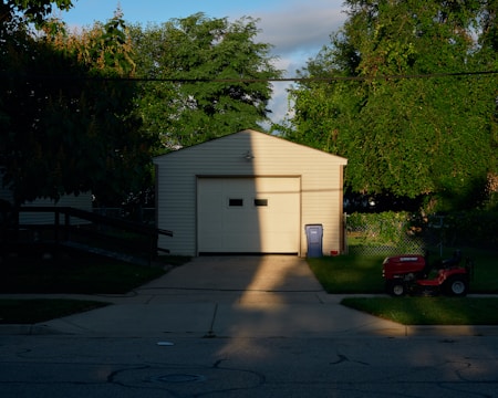 A garage with a closed door is partially bathed in sunlight, creating a stark shadow across the structure. Lush green trees surround the building, and the lawn in front hosts a red lawn mower. A blue recycling bin stands to the right of the garage on the edge of the driveway.