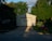 A garage with a closed door is partially bathed in sunlight, creating a stark shadow across the structure. Lush green trees surround the building, and the lawn in front hosts a red lawn mower. A blue recycling bin stands to the right of the garage on the edge of the driveway.