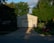 A garage with a closed door is partially bathed in sunlight, creating a stark shadow across the structure. Lush green trees surround the building, and the lawn in front hosts a red lawn mower. A blue recycling bin stands to the right of the garage on the edge of the driveway.