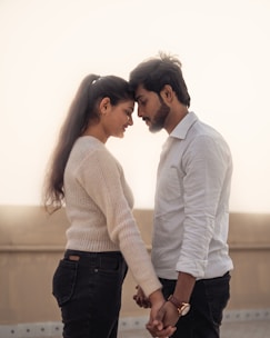 A couple stands closely together, gently holding hands and touching foreheads. The woman has long hair tied back and wears a light-colored sweater and dark jeans. The man has a short beard, styled hair, and wears a light-colored buttoned shirt with dark jeans. The background is softly blurred, providing a serene setting.