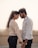A couple stands closely together, gently holding hands and touching foreheads. The woman has long hair tied back and wears a light-colored sweater and dark jeans. The man has a short beard, styled hair, and wears a light-colored buttoned shirt with dark jeans. The background is softly blurred, providing a serene setting.