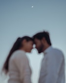 Silhouetted figures of two people standing close with their foreheads touching against a soft, gradient sky. A small crescent moon appears overhead, casting a serene atmosphere.