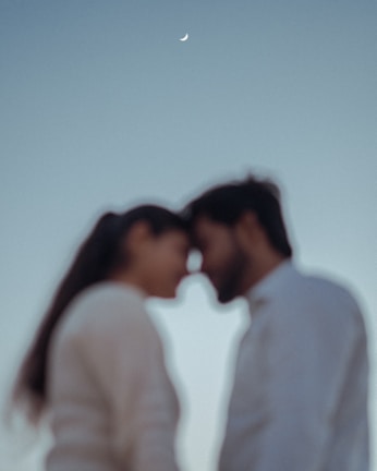 Silhouetted figures of two people standing close with their foreheads touching against a soft, gradient sky. A small crescent moon appears overhead, casting a serene atmosphere.