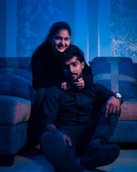 A couple is sitting together in a dimly lit, cozy room with blue lighting. The woman is sitting on a couch, leaning forward and hugging the man, who is seated on the floor, leaning against the couch. They are both dressed in dark clothing. The wallpaper in the background features a floral pattern, and there is a sense of intimacy and warmth in their posture and expressions.