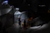 The image depicts a dimly lit exhibit with angular, geometric structures resembling a cave or bunker. Two people are observing posters and text on a wall, with a sign that includes Cyrillic text nearby. A stack of sandbags is visible, adding to the war-like or military atmosphere.