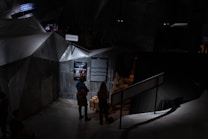 The image depicts a dimly lit exhibit with angular, geometric structures resembling a cave or bunker. Two people are observing posters and text on a wall, with a sign that includes Cyrillic text nearby. A stack of sandbags is visible, adding to the war-like or military atmosphere.