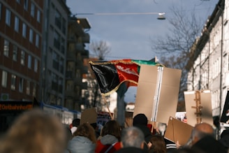 A group of people is gathered, marching along a city street. They are holding signs and banners, including a large, black, red, and green flag with a central emblem. The scene is captured in the daytime, with multi-story buildings lining the street, and a clear sky overhead.