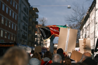 A group of people is gathered, marching along a city street. They are holding signs and banners, including a large, black, red, and green flag with a central emblem. The scene is captured in the daytime, with multi-story buildings lining the street, and a clear sky overhead.
