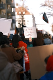 A group of people is participating in a protest or rally. One individual holds a sign that reads 'We stand with Afghan women.' The crowd appears to be moving together in an urban setting potentially signified by residential buildings and flags. The atmosphere suggests a display of solidarity.