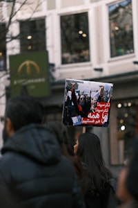 A group of people at a protest or demonstration is visible, with one individual holding up a placard featuring an image of a crowd holding signs and a message written in what appears to be Persian script. The background showcases a tall building with large windows, and the image has an overall urban setting.