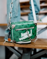 A green designer handbag with white lettering and a monogram pattern is resting on a wooden bench. It features a zipper and a silver chain strap with metal hardware. The background includes blurred denim-clad legs, suggesting a casual setting.