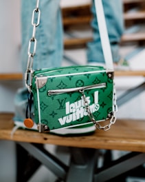 A green designer handbag with white lettering and a monogram pattern is resting on a wooden bench. It features a zipper and a silver chain strap with metal hardware. The background includes blurred denim-clad legs, suggesting a casual setting.