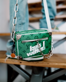A green designer handbag with white lettering and a monogram pattern is resting on a wooden bench. It features a zipper and a silver chain strap with metal hardware. The background includes blurred denim-clad legs, suggesting a casual setting.