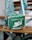 A green designer handbag with white lettering and a monogram pattern is resting on a wooden bench. It features a zipper and a silver chain strap with metal hardware. The background includes blurred denim-clad legs, suggesting a casual setting.