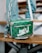 A green designer handbag with white lettering and a monogram pattern is resting on a wooden bench. It features a zipper and a silver chain strap with metal hardware. The background includes blurred denim-clad legs, suggesting a casual setting.