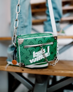 A green designer handbag with white lettering and a monogram pattern is resting on a wooden bench. It features a zipper and a silver chain strap with metal hardware. The background includes blurred denim-clad legs, suggesting a casual setting.