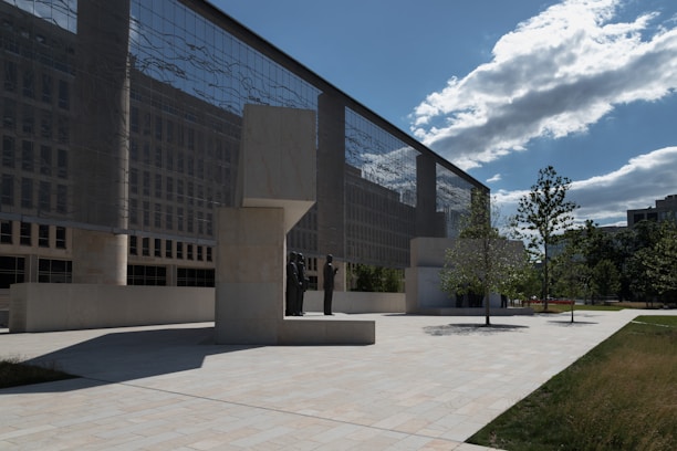 A modern architecture building with a reflective glass facade stands adjacent to an open courtyard. The courtyard consists of light stone pavement and is bordered by grass and trees. Several statues are sculpted under a structured canopy, casting shadows on the ground. The sky is partly cloudy with bright sunlight enhancing the contrast.