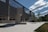 A modern architecture building with a reflective glass facade stands adjacent to an open courtyard. The courtyard consists of light stone pavement and is bordered by grass and trees. Several statues are sculpted under a structured canopy, casting shadows on the ground. The sky is partly cloudy with bright sunlight enhancing the contrast.