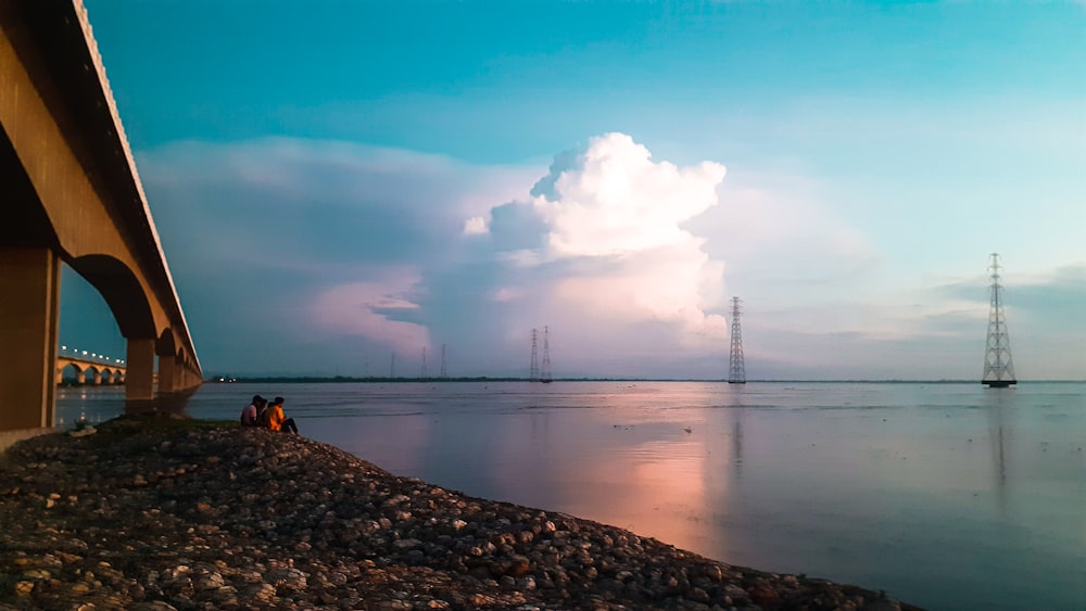 a couple of people sitting on a beach under a bridge