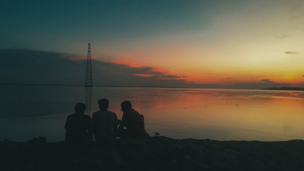 three people sitting on a beach watching the sunset