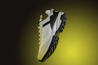 A modern, rugged sneaker is presented against a gradient backdrop that transitions from dark green to bright yellow. The shoe features a combination of materials, including white textured panels, black accents, and a robust black sole with heavy treading.
