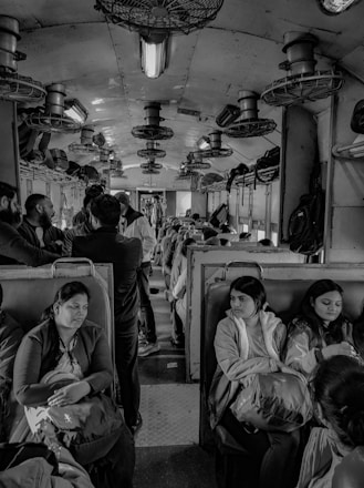 The interior of a crowded train carriage filled with passengers seated and standing. Overhead fans and luggage racks are visible, creating a busy and somewhat cramped environment.