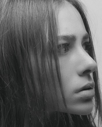 A close-up profile shot of a person's face, focusing on the eye, nose, and lips. The image is in black and white, highlighting the texture of the skin and the strands of hair falling across the face.
