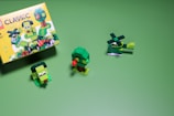 A set of small, green LEGO creations is arranged on a green background. The box on the left features colorful illustrations of more LEGO builds. On the right, a helicopter, a tree, and a small green character with eyes are made from LEGO bricks.
