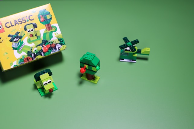 A set of small, green LEGO creations is arranged on a green background. The box on the left features colorful illustrations of more LEGO builds. On the right, a helicopter, a tree, and a small green character with eyes are made from LEGO bricks.