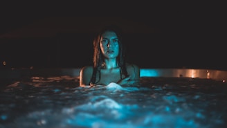 A woman with wet hair stands in a hot tub filled with bubbling water. The lighting is dark with a cool blue and teal tone, creating a mysterious and moody atmosphere.
