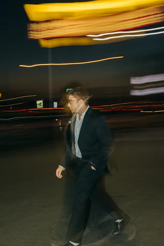 A man in a tailored, unconventional formal coat walking through a city street at dusk.
