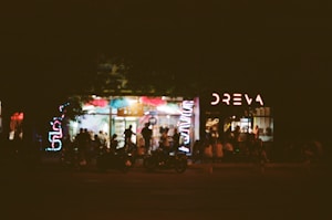 A dimly lit outdoor scene featuring a bustling caf&eacute; or bar with neon lights outside. People are gathered in small groups, socializing near the entrance. The atmosphere suggests a lively and vibrant night setting.