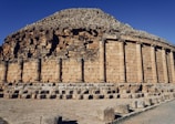 A large, ancient stone structure featuring a circular arrangement with tall, cylindrical columns. The stones are weathered and have a light brown color, indicative of historical architecture. The sky above is clear, accentuating the structure's grandeur.