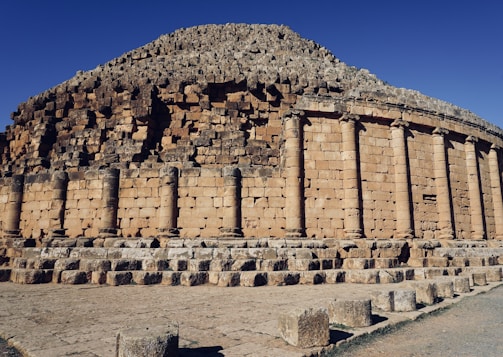 A large, ancient stone structure featuring a circular arrangement with tall, cylindrical columns. The stones are weathered and have a light brown color, indicative of historical architecture. The sky above is clear, accentuating the structure's grandeur.