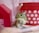 A small rodent is standing on its hind legs against a background with a red plastic container adorned with white heart patterns and a textured pink fabric. There is also a red toy house structure on the left side.