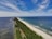A long, narrow peninsula bordered by a sandy beach on one side, extending into the sea. Dense green vegetation covers the land, with a road running alongside it. The sky is filled with wispy clouds and the ocean water is a mix of deep blue and lighter shades near the shore.