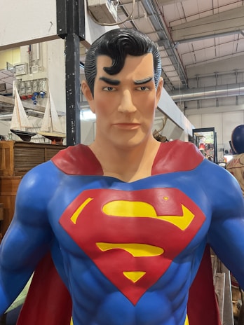A large, detailed statue of a superhero figure is prominently displayed, featuring a muscular build and iconic costume. The suit is bright blue with a large red and yellow emblem on the chest, resembling a stylized letter, and a red cape draped over the shoulders. The statue has a confident expression, meticulously styled black hair, and a backdrop of industrial elements, including sailboat models and furniture.