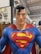 A large, detailed statue of a superhero figure is prominently displayed, featuring a muscular build and iconic costume. The suit is bright blue with a large red and yellow emblem on the chest, resembling a stylized letter, and a red cape draped over the shoulders. The statue has a confident expression, meticulously styled black hair, and a backdrop of industrial elements, including sailboat models and furniture.