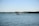 A motorboat is moving across a calm body of water with gentle waves. The background features a distant shoreline with several hills and some scattered houses visible along the coast under a clear blue sky.