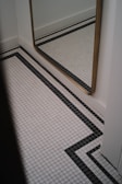 A corner of a room featuring a large mirror with a golden frame mounted on a white wall. The floor is tiled with a white grid pattern, accented by black lines forming geometric shapes.